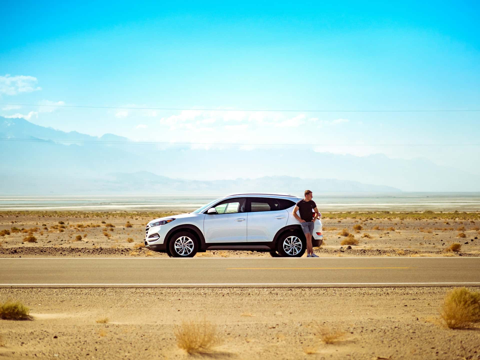 A white SUV stopped on the gravel shoulder of an open highway in bright daylight, with the driver standing at the rear of the vehicle