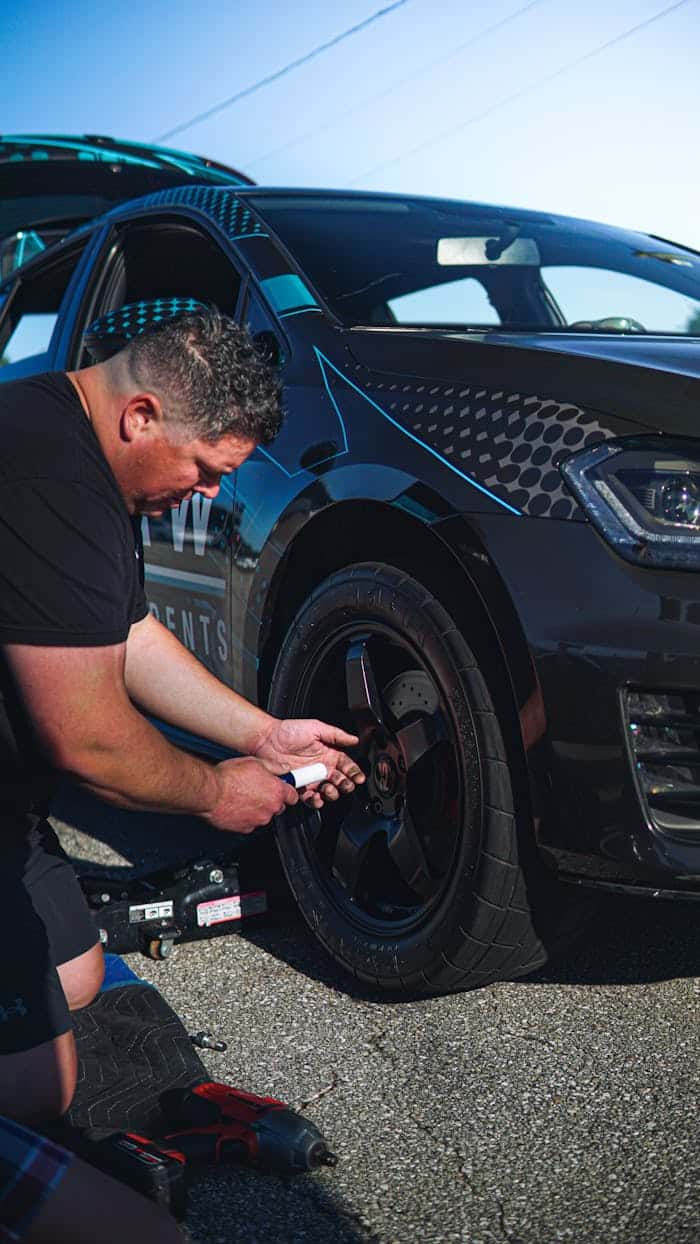 A technician kneeling on the asphalt beside a black hatchback, fitting a wheel back on with a trolley jack and impact wrench laid out on the road
